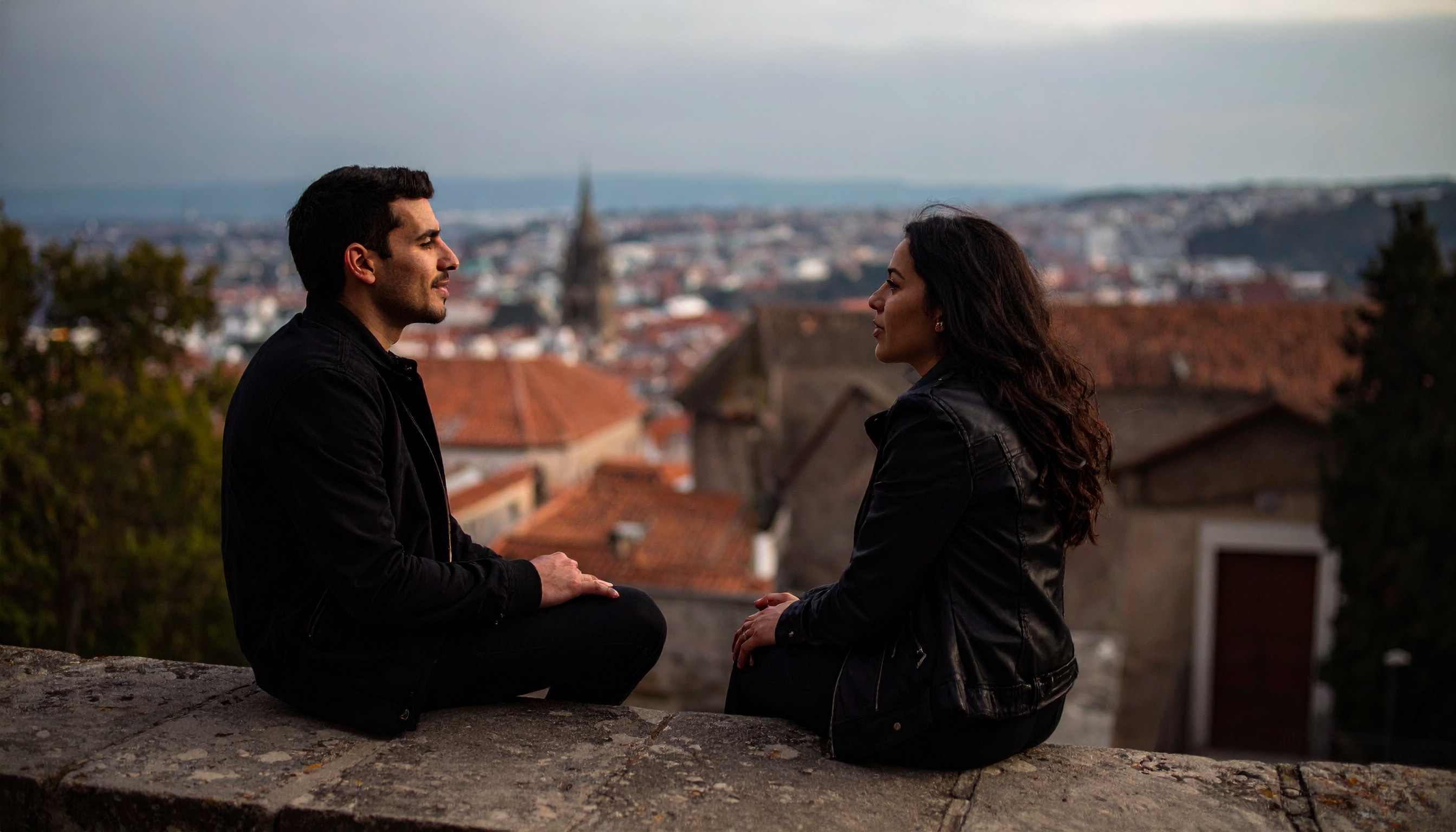 Ein Mann und eine Frau sitzen einander gegenüber auf einer Mauer und sprechen miteinander, im Hintergrund rote Dächer einer Altstadt.