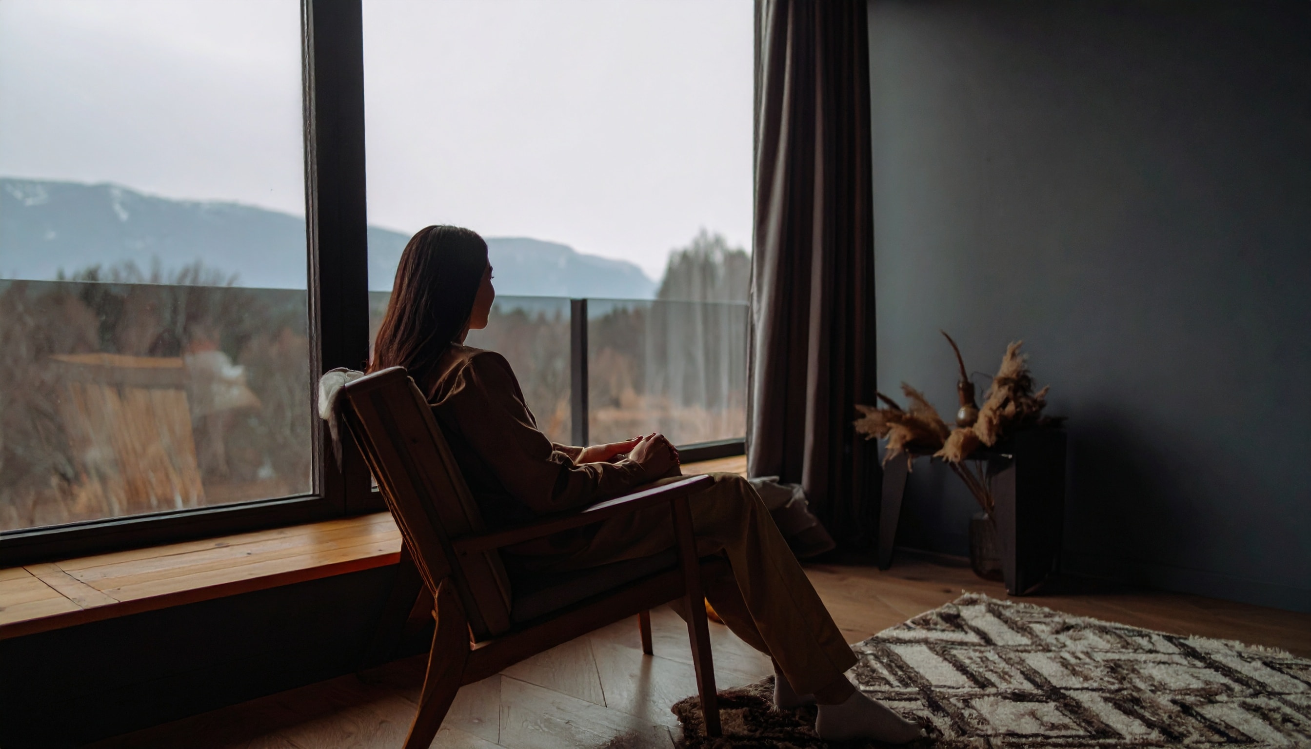 Eine Frau sitzt in einem Holzsessel am Fenster und schaut hinaus auf eine Berglandschaft.